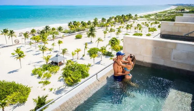 Two people embrace in an infinity pool overlooking a sandy beach with palm trees and turquoise water