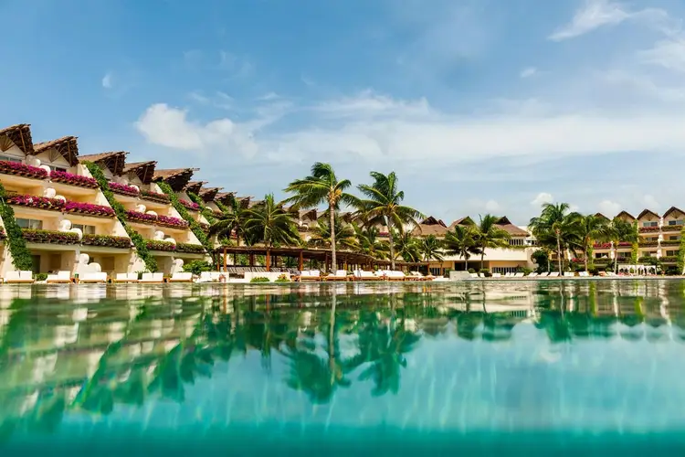 Beachfront resort buildings with tiered terraces and flowering vines overlook clear turquoise water lined with palm trees
