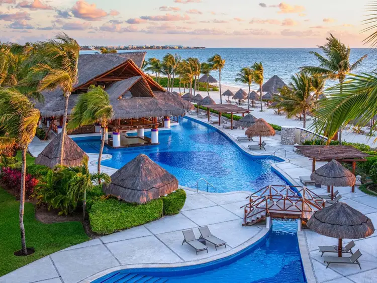 Aerial view of a beachfront resort with a large swimming pool, thatched-roof structures, palm trees, and ocean beyond at sunset