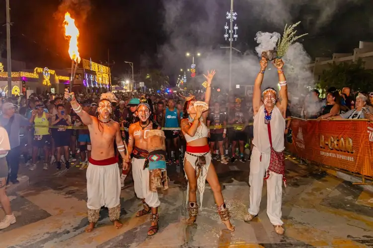 Performers in traditional dress and face paint dance in the street at night surrounded by crowds and ceremonial fire