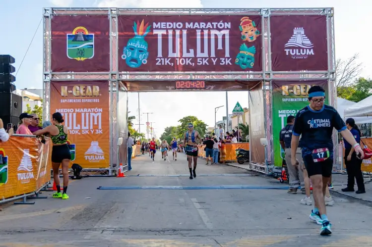 Runners cross the finish line at a half marathon race under a large red and white banner displaying race information and distance options
