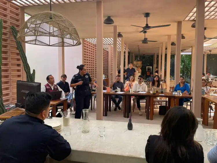 A woman stands to address a group of people seated at tables in an open-air covered venue during what appears to be a meeting or forum