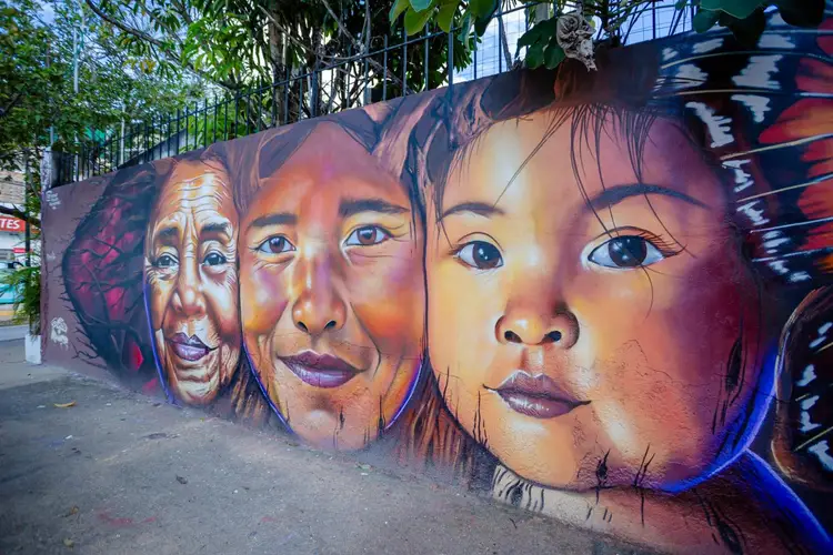 Mural depicting four painted faces of varying ages in warm and cool tones on an urban street wall beneath overhanging vegetation