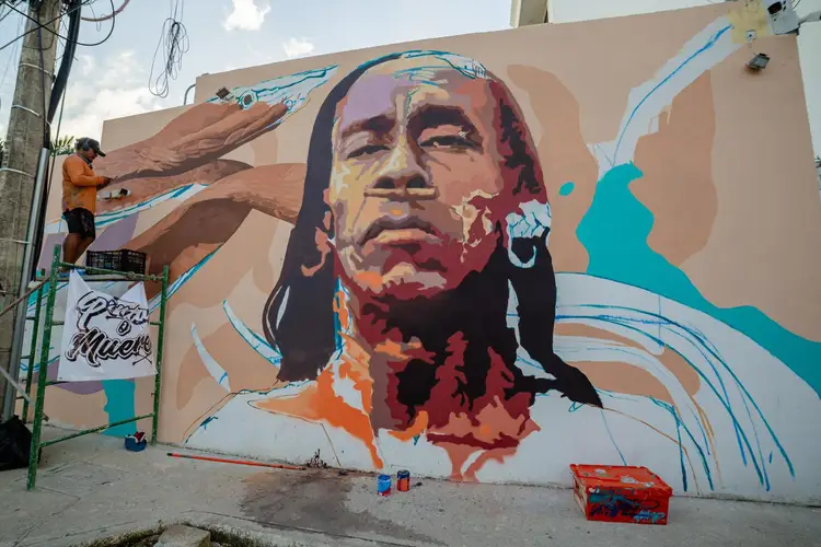 Street artist on scaffolding works on a large mural featuring a portrait with flowing hair and colorful abstract elements on a building wall