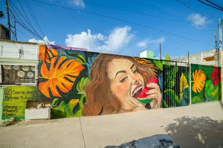 Large mural on a building wall depicts a woman with brown hair eating watermelon, surrounded by colorful orange and yellow flowers against a green background