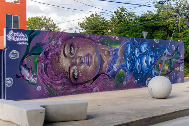 A large mural painted on a dark blue wall depicts a face with purple hair and surreal imagery, surrounded by concrete bollards and an orange building, along a s