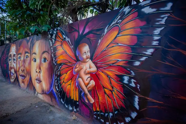 Mural depicting human faces and a figure with large butterfly wings in orange, red and purple tones on a street wall beneath palm trees