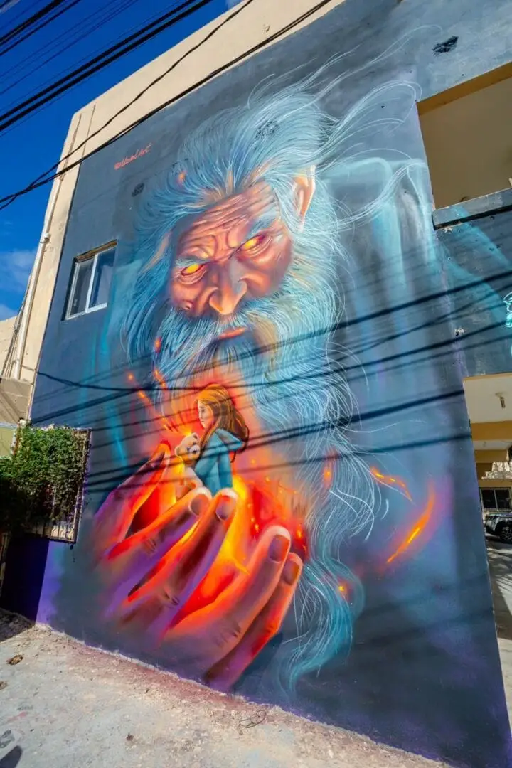 Mural of an elderly figure with white hair and glowing eyes holding flames, painted on a building wall in an urban setting