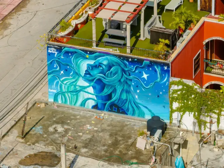 Large blue mural depicting a woman's face with flowing hair and stars covers the side of a building in an urban courtyard setting