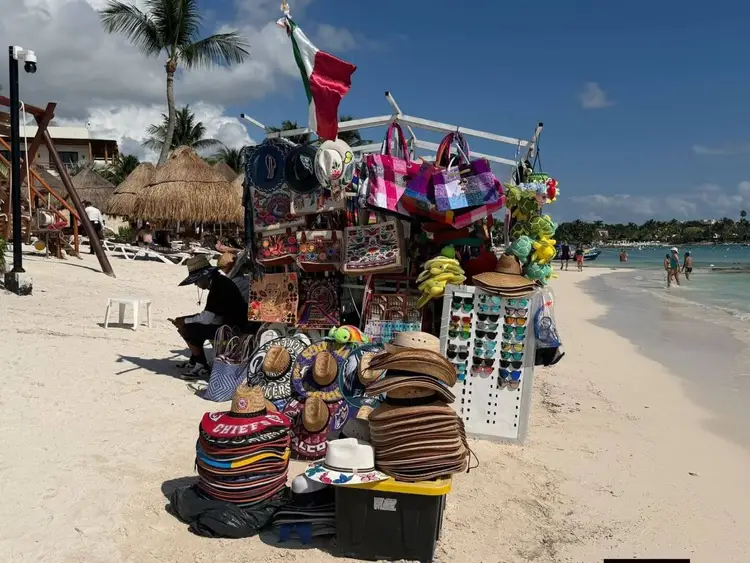 A beach vendor's stall displays colorful hats, bags, and souvenirs on sand near the water's edge, with beachgoers and palm-thatched structures visible in the ba A beach vendor's stall displays colorful hats, bags, and souvenirs on sand near the water's edge, with beachgoers and palm-thatched structures visible in the ba