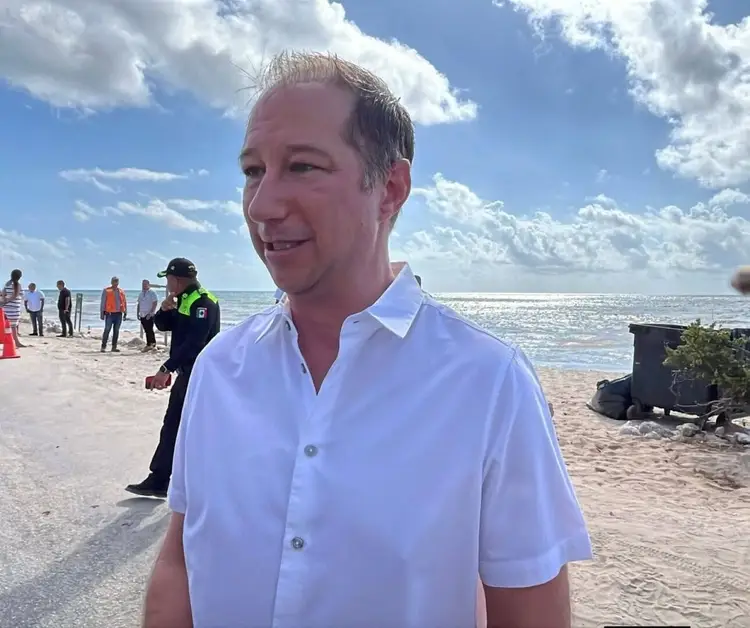 Man in white button-up shirt smiles at camera on a sandy beach with ocean and other people visible in the background Man in white button-up shirt smiles at camera on a sandy beach with ocean and other people visible in the background