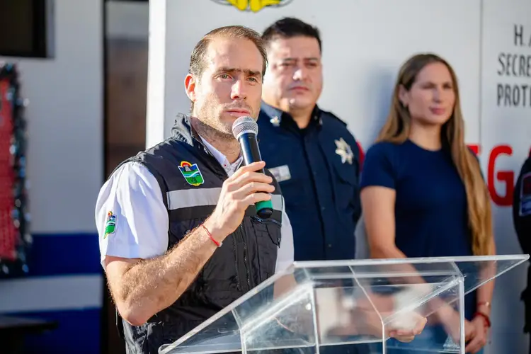 Man in police uniform speaking at a podium with microphone, flanked by two other officials in uniform at an indoor event
