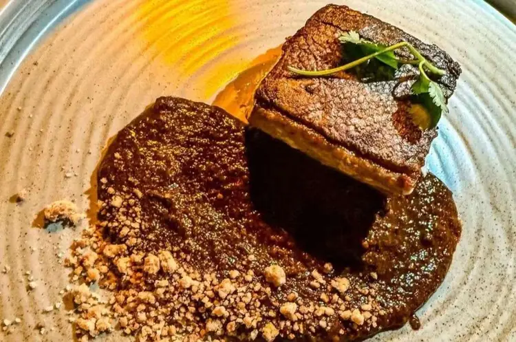 A plated dessert featuring a chocolate cake slice dusted with cocoa powder, surrounded by crumble and a drizzle of yellow sauce, garnished with green herbs A plated dessert featuring a chocolate cake slice dusted with cocoa powder, surrounded by crumble and a drizzle of yellow sauce, garnished with green herbs