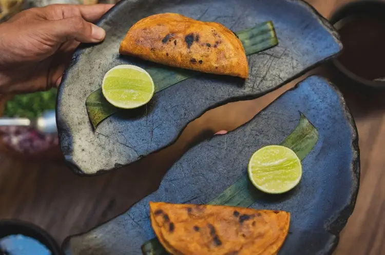 Person holds a blue fish-shaped plate with two fried pastries, lime halves, and green garnish arranged on it Person holds a blue fish-shaped plate with two fried pastries, lime halves, and green garnish arranged on it