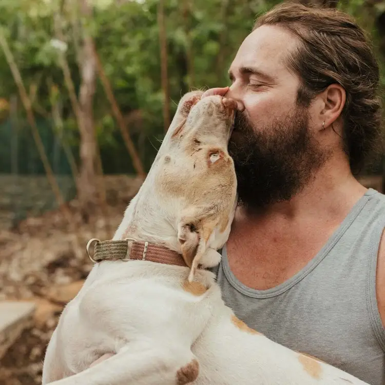 Man with beard holding a tan and white dog that is licking his face outdoors near trees