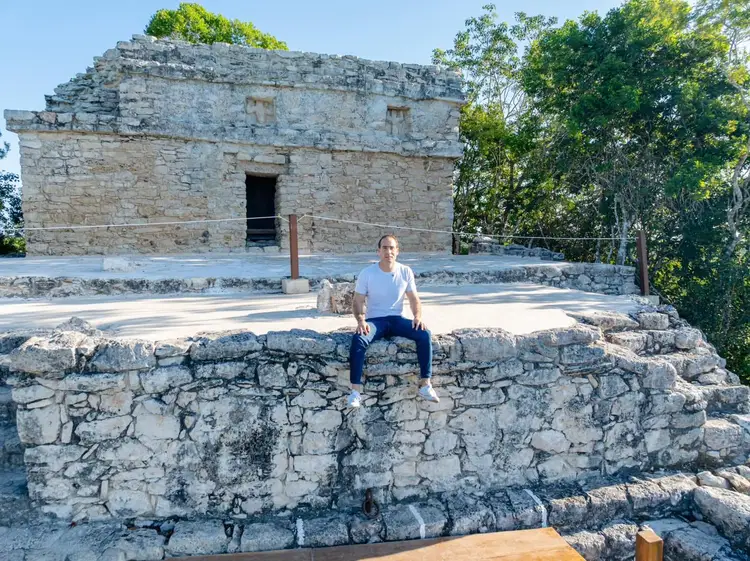 Man sitting on ancient stone ruins in front of a weathered stone structure with an entrance doorway and surrounding trees