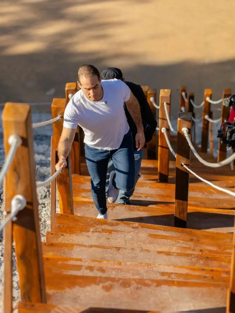Man walking down a wooden staircase with rope railings while carrying a child on his back