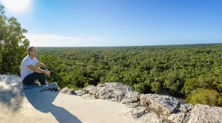 Man in white shirt crouching on rocky outcrop overlooking dense forest canopy under clear sky