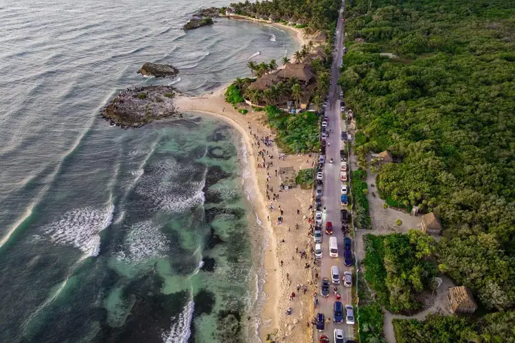 Aerial view of a crowded beach with parked cars along a coastal road, dense forest to the right, and rocky outcrops in the shallow water