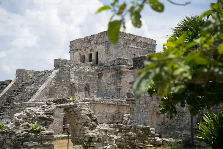 Ancient stone ruins with stepped pyramid structures and a rectangular tower stand among overgrown vegetation under a cloudy sky Ancient stone ruins with stepped pyramid structures and a rectangular tower stand among overgrown vegetation under a cloudy sky