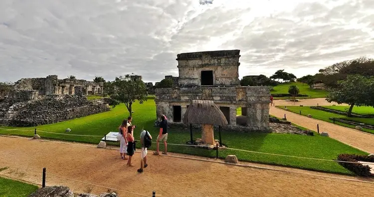 Visitors walk through an archaeological site with stone structures and ruins surrounded by green lawns and trees under a cloudy sky Visitors walk through an archaeological site with stone structures and ruins surrounded by green lawns and trees under a cloudy sky