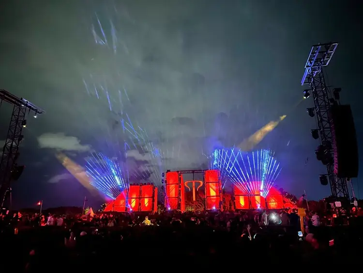 Large illuminated stage with red panels and blue laser light displays at an outdoor concert venue with crowd silhouettes in foreground and tall lighting rigs on Large illuminated stage with red panels and blue laser light displays at an outdoor concert venue with crowd silhouettes in foreground and tall lighting rigs on