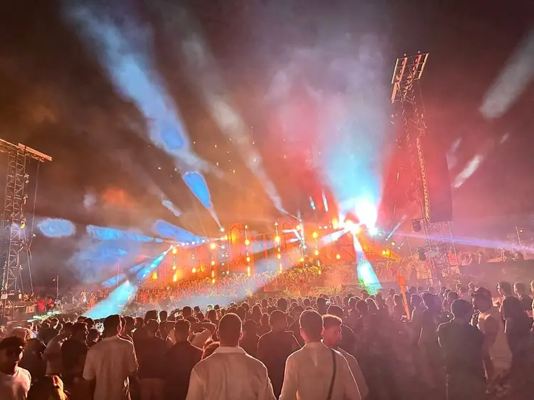 Large crowd of attendees facing an outdoor stage with dramatic lighting effects and tall towers at night Large crowd of attendees facing an outdoor stage with dramatic lighting effects and tall towers at night