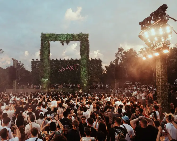 Large crowd of people gather in an outdoor venue with a massive ivy-covered stage structure and illuminated lighting rigs overhead