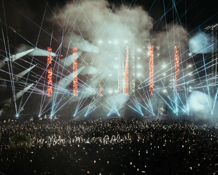 Large crowd watches an outdoor concert stage with illuminated red vertical structures, blue laser beams, and fog effects at night