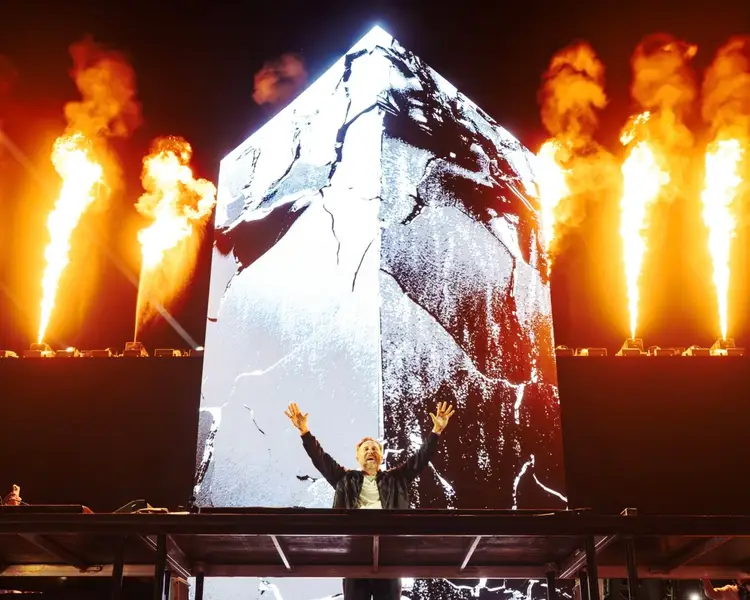 Person on stage with arm raised beneath large projection screen, surrounded by towering flames shooting upward into the night sky