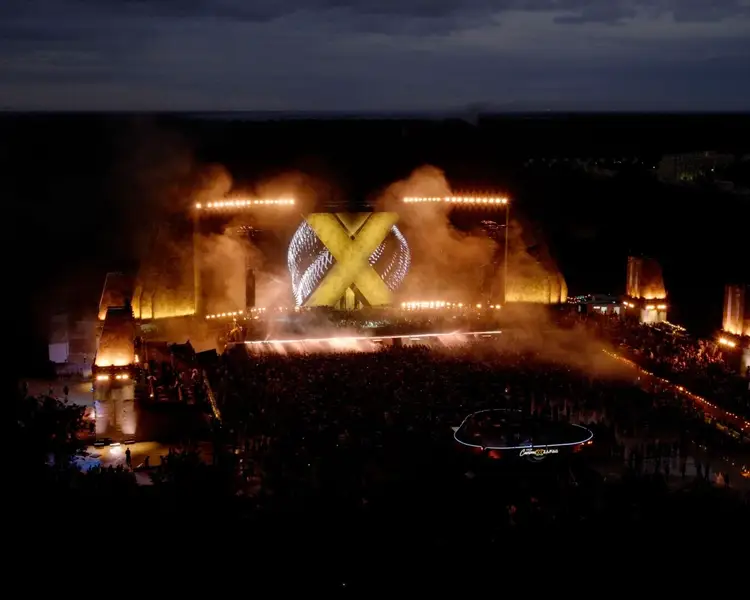 Large outdoor music festival stage with illuminated X logo and surrounding lights, crowd gathered in front at night