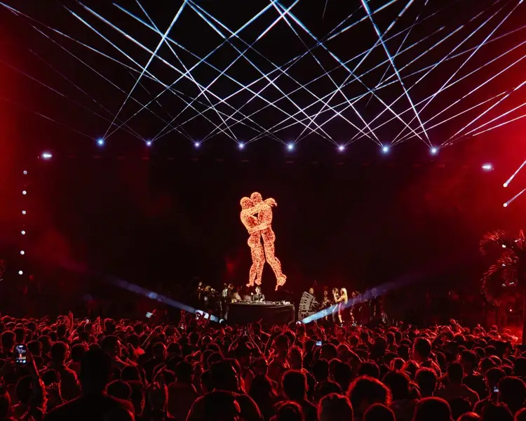A large crowd watches a glowing bear figure projected on stage during an electronic music concert with red lighting and laser effects overhead