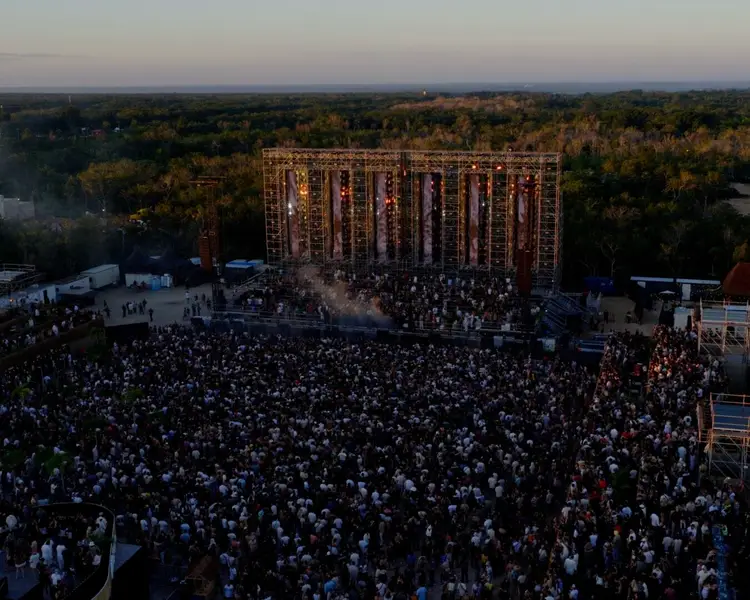 Aerial view of a large outdoor music festival with thousands of attendees gathered in front of a massive stage structure surrounded by forested landscape at dus