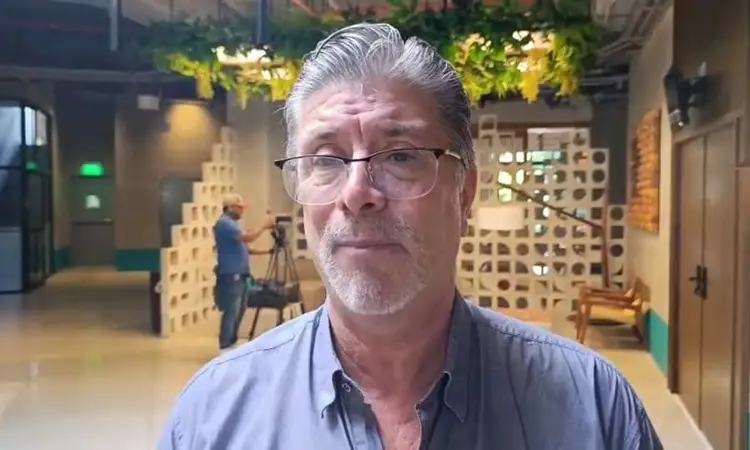 Man wearing glasses and blue shirt stands in an interior space with wooden block installations and another person visible in the background Man wearing glasses and blue shirt stands in an interior space with wooden block installations and another person visible in the background