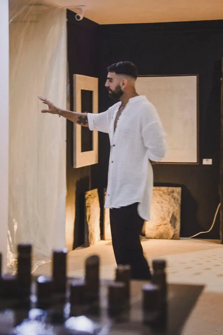 Man in white shirt gestures toward framed artwork displayed on a gallery wall during an exhibition