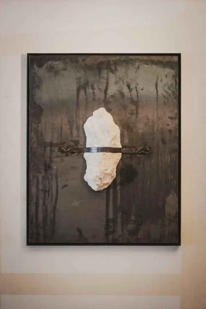 Framed artwork displaying a white stone or mineral specimen pierced by a metal rod against a darkened, textured background