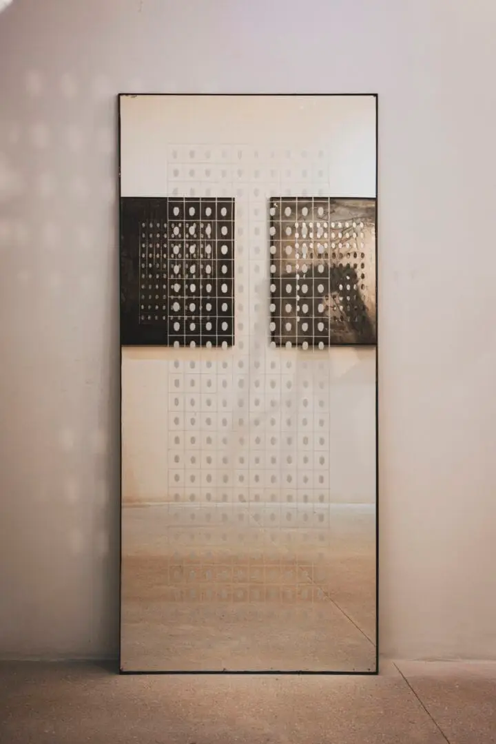 A tall rectangular mirror leans against a white wall, reflecting perforated metal panels and architectural elements of an interior space