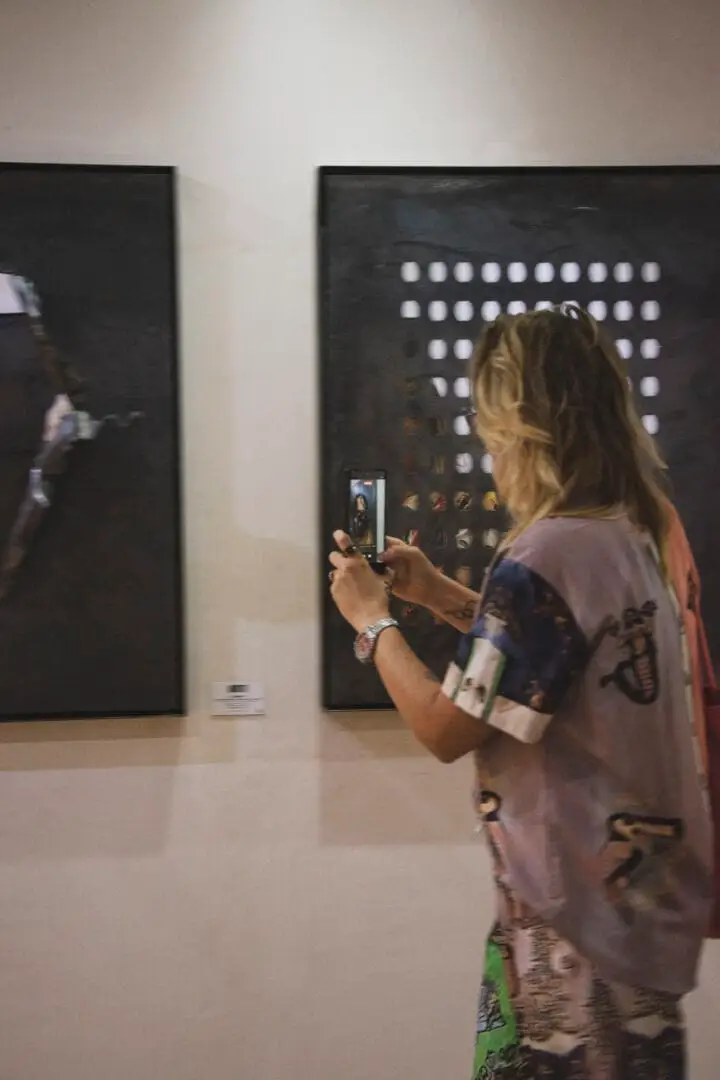 Woman photographs a dark artwork displaying a grid of illuminated white squares on a gallery wall