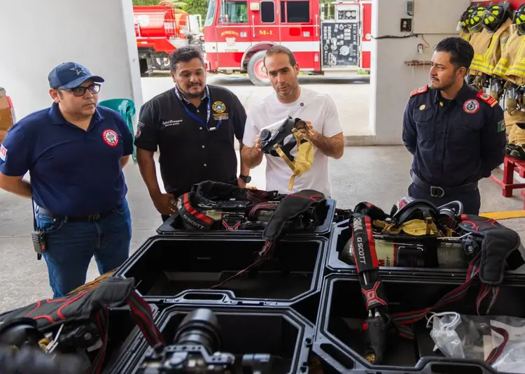 Four firefighters examine rescue equipment and gear laid out on black cases in a fire station bay Four firefighters examine rescue equipment and gear laid out on black cases in a fire station bay