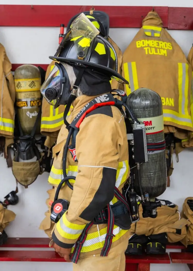 Firefighter gear hangs on display, including a tan turnout suit with reflective stripes, black helmet with face shield, and air tank labeled with 3M branding Firefighter gear hangs on display, including a tan turnout suit with reflective stripes, black helmet with face shield, and air tank labeled with 3M branding
