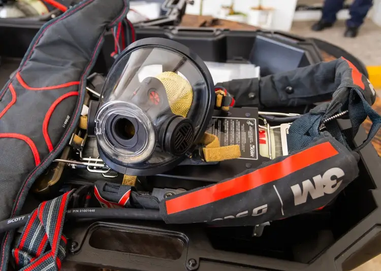 A full-face respirator mask with yellow filters sits in an open gear bag alongside protective equipment and straps A full-face respirator mask with yellow filters sits in an open gear bag alongside protective equipment and straps