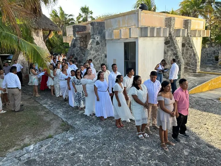 A crowd of people in white and pastel clothing gather outside a concrete building surrounded by palm trees in a tropical setting A crowd of people in white and pastel clothing gather outside a concrete building surrounded by palm trees in a tropical setting