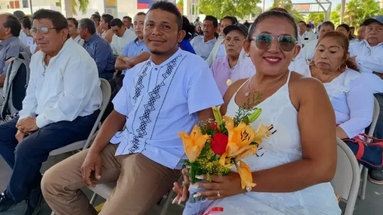 Woman holding a bouquet of flowers sits beside a man wearing a decorative shirt at an outdoor community gathering with many people in attendance Woman holding a bouquet of flowers sits beside a man wearing a decorative shirt at an outdoor community gathering with many people in attendance