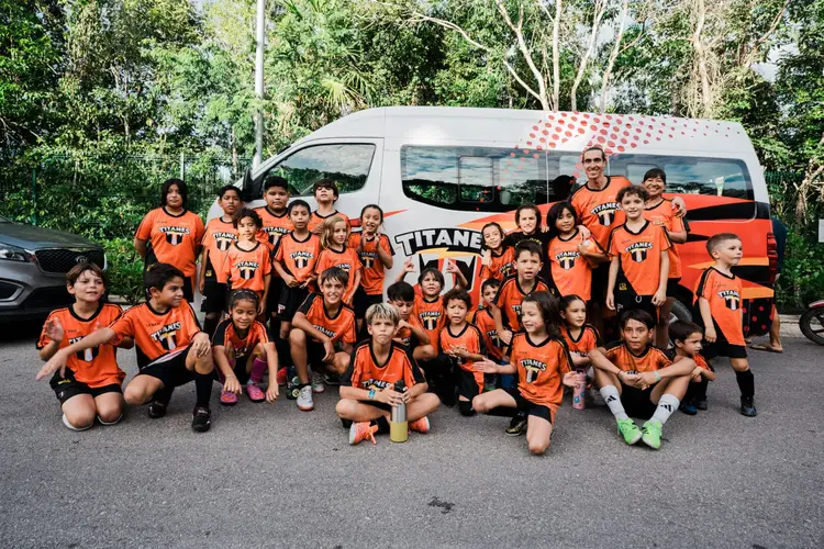 Large group of children wearing orange sports uniforms pose together in front of a white van with orange stripe and "Titans" text in a parking lot surrounded by Large group of children wearing orange sports uniforms pose together in front of a white van with orange stripe and "Titans" text in a parking lot surrounded by