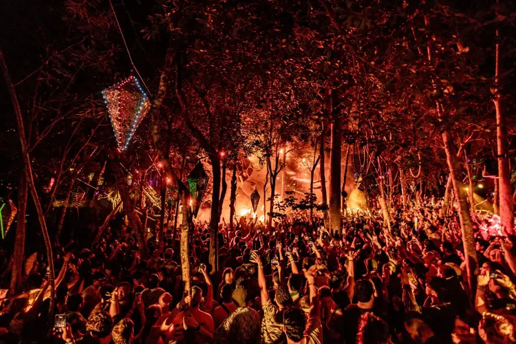 Large crowd of people gathered in a tree-filled outdoor venue illuminated by warm orange and red lighting at night Large crowd of people gathered in a tree-filled outdoor venue illuminated by warm orange and red lighting at night