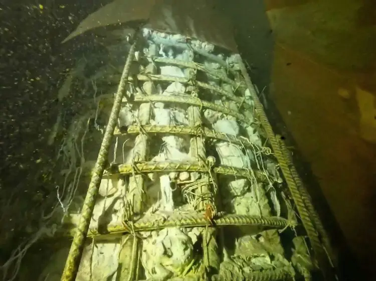 Underwater view of a corroded metal ladder or structural framework covered in sediment and marine growth on what appears to be a sunken vessel or submerged infr Underwater view of a corroded metal ladder or structural framework covered in sediment and marine growth on what appears to be a sunken vessel or submerged infr