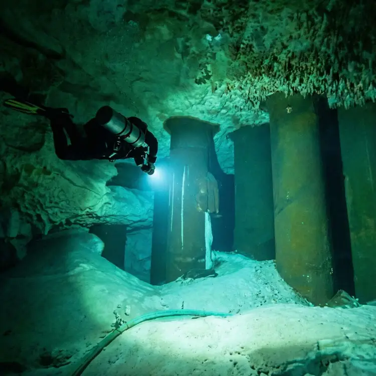 A scuba diver with a headlamp explores an underwater cavern with metal pillars and sandy bottom A scuba diver with a headlamp explores an underwater cavern with metal pillars and sandy bottom