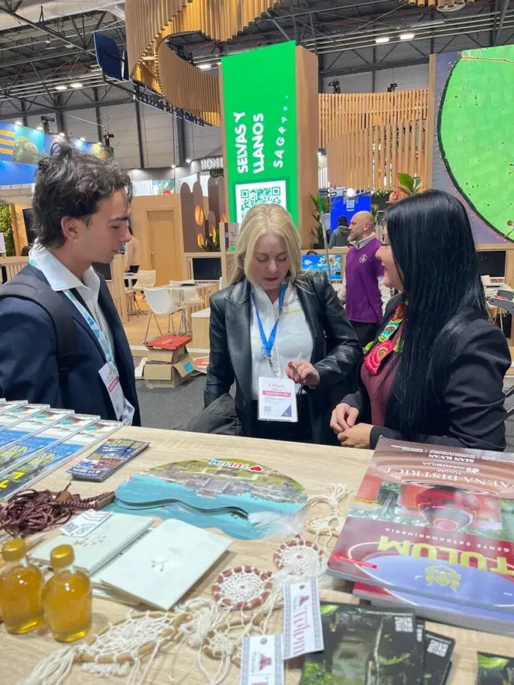 Three people wearing credential badges examine tourism materials and products at a convention booth featuring wooden design elements and a green digital display