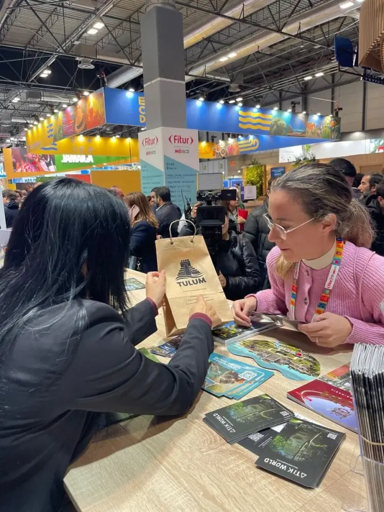 Visitors receive promotional materials at a tourism trade show booth displaying colorful signage and travel brochures inside a large convention hall