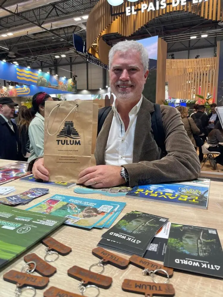 Man holds a paper bag with a Tulum logo at an indoor tourism convention booth displaying promotional materials and wooden keychains